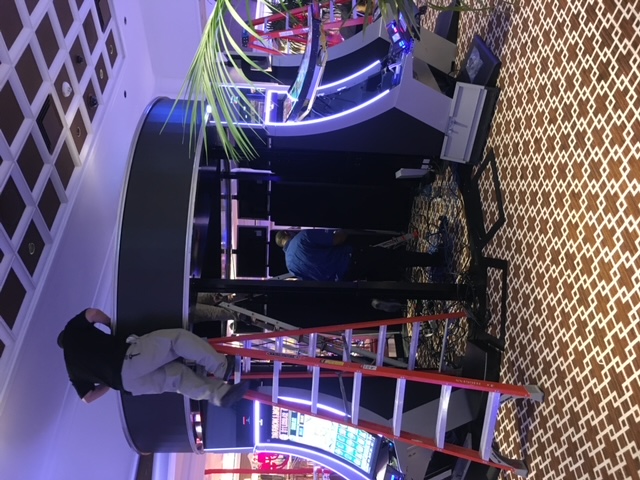 Technicians assembling a large circular overhead gaming display between slot machines.